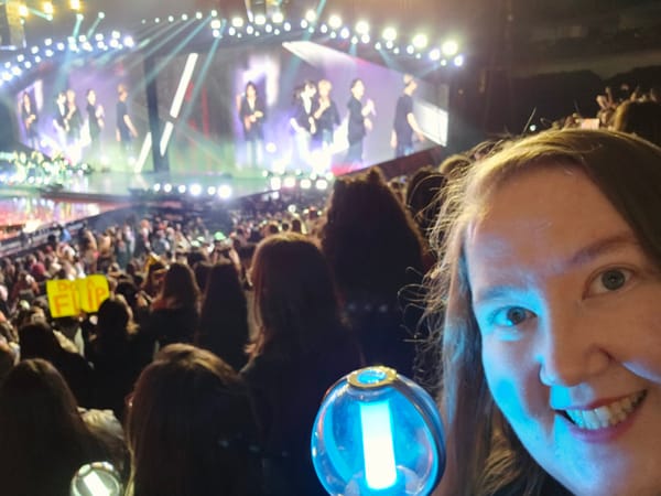 Mari holding a light stick in front of a stadium of people with the ENHYPEN band on stage.